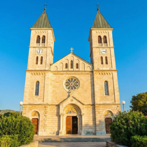 Front kościoła św. Jakuba w Medjugorie z jego dwiema charakterystycznymi wieżami, skąpany w promieniach słońca na tle głębokiego błękitu nieba.
